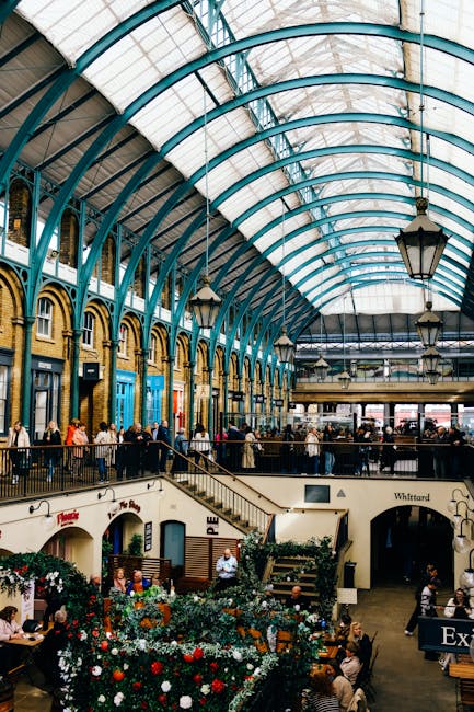 The image depicts the interior of a bustling market hall with a high arched glass ceiling supported by teal-colored steel framework. Natural light floods the space, illuminating customers browsing and
