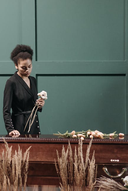A young woman with dark curly hair, dressed in a black blazer with metallic button details, stands gracefully by a wooden funeral arrangement table. She holds a small white flower in her right hand, a