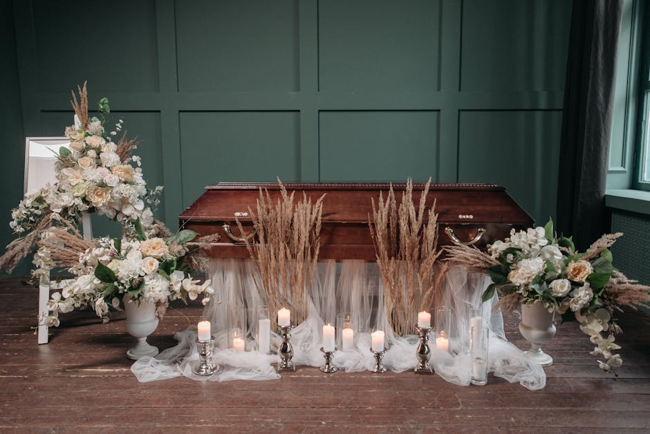 A funeral floral arrangement displayed in front of a vintage wooden casket with an elegant dark green paneled wall background. The arrangement includes two large white ceramic vases filled with soft c