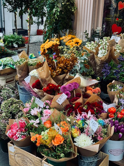 A vibrant display of fresh flower bouquets arranged outdoors on a table, featuring a variety of blooms including bright yellow sunflowers, pink and white lilies, red and purple chrysanthemums, orange 